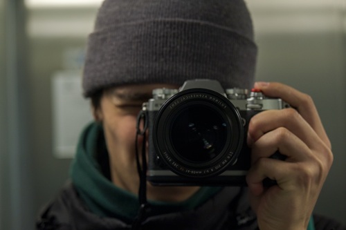 Person wearing a gray beanie and green jacket holding a camera up to take a photo in what appears to be an indoor setting with soft lighting and a calm, neutral atmosphere